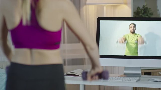 Rear view of woman doing front and lateral dumbbell raises at home and watching video on computer with online personal trainer