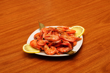 Freshly prepared shrimps lie on a white plate, adorned with spices and lemon. Against the background of a wooden table.