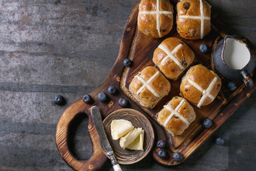 Hot cross buns on wooden cutting board served with butter, knife, fresh blueberries and jug of...