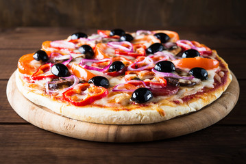 Italian pizza with salami, mushroom, tomatoes, sweet pepper, black olives and purple onion on dark wooden background close up. Italian traditional food. Popular street food.