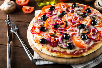 Italian pizza with salami, mushroom, tomatoes, sweet pepper, black olives and purple onion on dark wooden background close up. Italian traditional food. Popular street food.