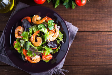 Fresh salad plate with shrimp, tomato and mixed greens (arugula, mesclun, mache) on wooden background top view. Healthy food. Clean eating.