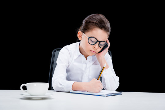 Tired little girl in formal wear using smartphone and taking notes