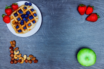 Healthy food. Green apple, nuts (cashew, hazel-nut, almond) and ceramic plate with gaufre and berries (blueberries, strawberries) isolated on wooden background