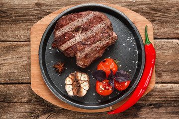 Appetizing beef steak decorated with grilled garlic, tomatoes, basil and chili pepper, on wooden rustic table. American food. Flat lay.