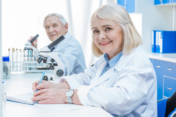 smiling senior chemists with microscope writing notes in laboratory
