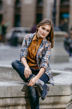 Fashion girl is sitting outdoors wearing blue jeans,brown checkered jacket and holding a brown handbag, urban city