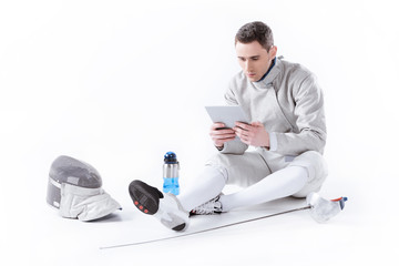 fencer in uniform using digital tablet while resting after training on white