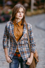 Fashion girl is walking on the sidewalk wearing blue jeans,brown checkered jacket and holding a brown handbag, urban city