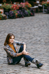 Fashion girl is sitting outdoors on the old cobblestone street wearing blue jeans,brown checkered jacket and holding a brown handbag, urban city