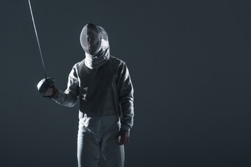 Professional fencer in fencing mask with rapier standing in position on grey