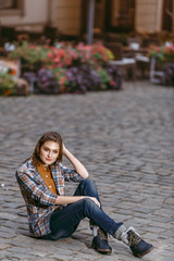 Fashion girl is sitting outdoors on the old cobblestone street wearing blue jeans,brown checkered jacket and holding a brown handbag, urban city