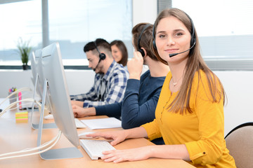 portrait of beautiful and cheerful young woman telephone operator with headset working on desktop computer in row in customer service call support helpline business center - obrazy, fototapety, plakaty