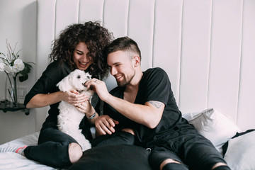 Man and woman in black play with little white dog on bed