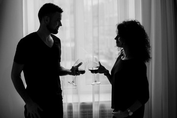 Man and woman clang glasses with wine standing before the window