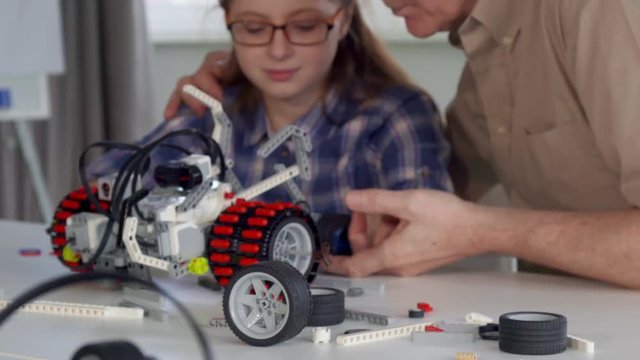 Senior caucasian man showing his granddaughter something on toy vehicle. Close up of different pieces of construction set on the table. Camera tilting up from the table with some part of toy car on it