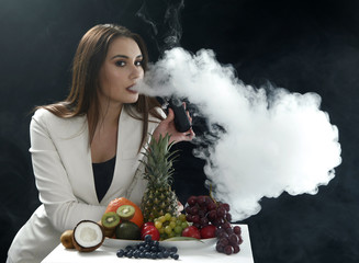 Young sexy girl smoking the vaping e cigarette close to assorti of differen fruits on black background