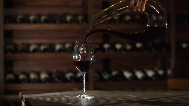The woman`s hand is pouring the red wine from the decanter in almost full wineglass at the backgroud of the wine warehouse.