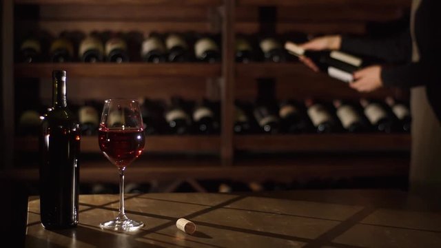 The cork, bottle and glass with red wine are placed on the table while woman is choosing the wine in the wine warehause.