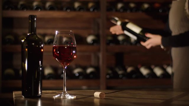 The composition of the bottle, cork and glass with red wine is placed on the wooden table while at the blurred background the sommelier is taking the wine, observing it and putting it back.