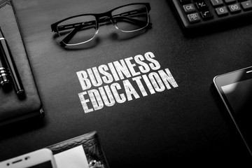 Black chalkboard with business accessories (notepad, magnifying glass, fountain pen, tablet, phone, glasses and calculator) and text BUSINESS EDUCATION. Top view.