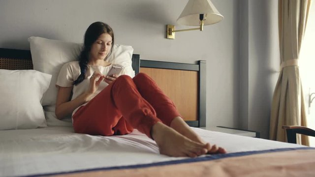 Pretty blonde woman lying in bed in hotel room and use smartphone and smiling