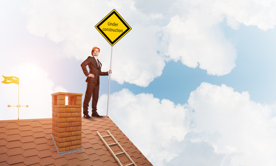 Young businessman on house brick roof holding yellow signboard. Mixed media