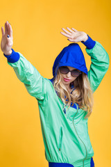 portrait of beautiful blond woman in sunglasses and blue green hooded jacket on yellow background. hipster summer.