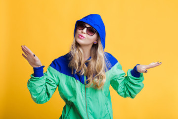 portrait of beautiful blond woman in sunglasses and blue green hooded jacket on yellow background. hipster summer.