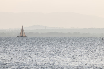 A small sailboat on a lake, with distant hills in the background, and very soft colors, mostly...