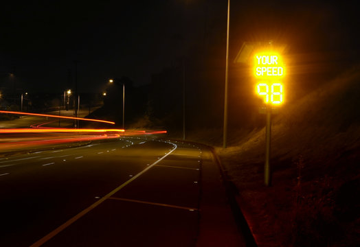 Streaks of taillights at night past a radar speed sign that indicates an excessive speed of 98 MPH