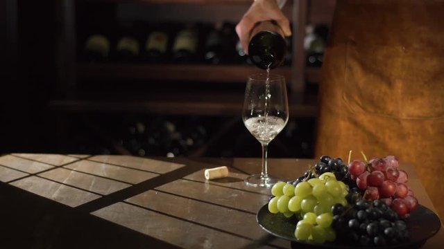 Close-up front portrait of the sommelier pouring the white wine from the bottle into the glass near the plate full of colourful grapes. The sun light on the table.
