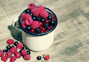 Fresh juicy berries, raspberries, currants, blackberries in an old white iron mug on a wooden surface, close up
