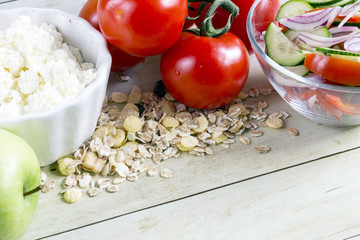 Foods for healthy eating: cottage chesse, tomatoes, apple, cereal, salad on light wood background