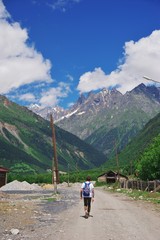 young man with a backpack walking along road past a village of buildings on the background of beautiful mountain scenery. On mountain tops there is snow on mountain slopes grow green forest.
