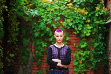 Beautiful punk girl with a purple mohawk standing against a background of brick wall overgrown with leaves.