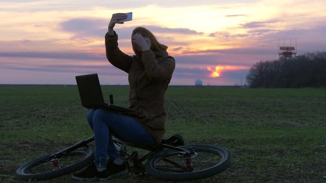 Smart red-haired girl uses laptop and makes photos near radar and communication equipment station in field during sunset with strong wind. Busy IT manager near airport on bicycle