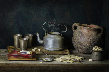 Classic still life with vintage tea kettle placed with silver cups,pottery jar,sugar,cigarettes box on rustic wooden background. © dinasaeed
