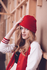 Stylish hipster woman in baseball jacket and red hat posing at wooden wall