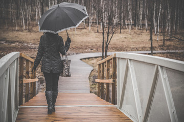 Young woman alone with umbrella walking on the footbridge. Gloomy atmosphere in the park. Cold, rainy spring day.