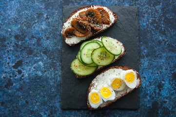 Mix of rye bread bruschettas with various filling. Mushrooms Quail Eggs Cucumber sandwiches served on a stone board on blue background. Flat lay.