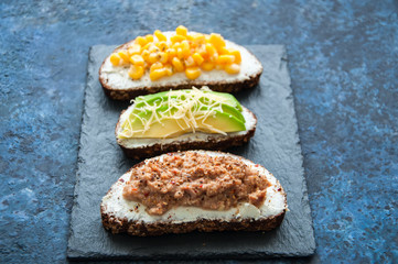 Mix of rye bread bruschettas with various filling. Sweet corn Omelette Tuna Seeds Cheese Avocado sandwiches served on a stone board on blue background. Flat lay.