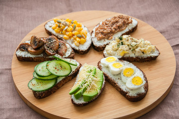 Mix of rye bread bruschettas with various filling. Sweet corn Avocado Mushrooms Quail Eggs Cucumber Omlette Tuna Seeds Cheese sandwiches served on a wooden round board. Flat lay.