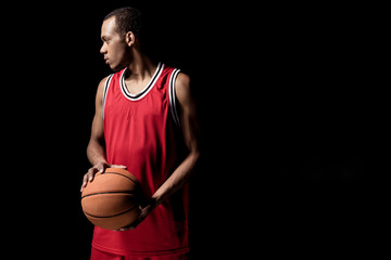 Young basketball player in uniform holding ball and looking away on black