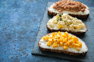 Mix of rye bread bruschettas with various filling. Sweet corn Omelette Tuna Seeds Cheese Avocado sandwiches served on a stone board on blue background. Flat lay.