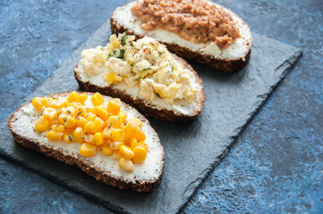 Mix of rye bread bruschettas with various filling. Sweet corn Omelette Tuna Seeds Cheese Avocado sandwiches served on a stone board on blue background. Flat lay.