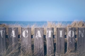 closeup of word vacation written on wooden fence with in front of grass covered dunes and blue sea...