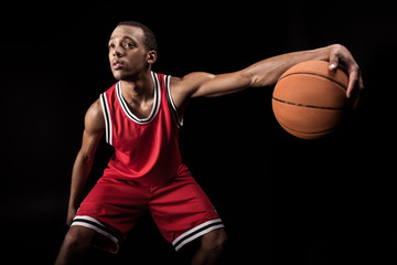 african sporty man in sports uniform playing basketball on black