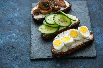 Mix of rye bread bruschettas with various filling. Mushrooms Quail Eggs Cucumber sandwiches served on a stone board on blue background. Flat lay.