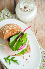 Sandwich with salad herbs chicken ham red onion seeds and spicy sour cream sauce served on a white plate. Wooden background.